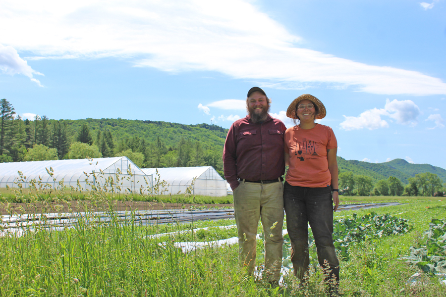 Two people smile in front of crop fields and hoop houses