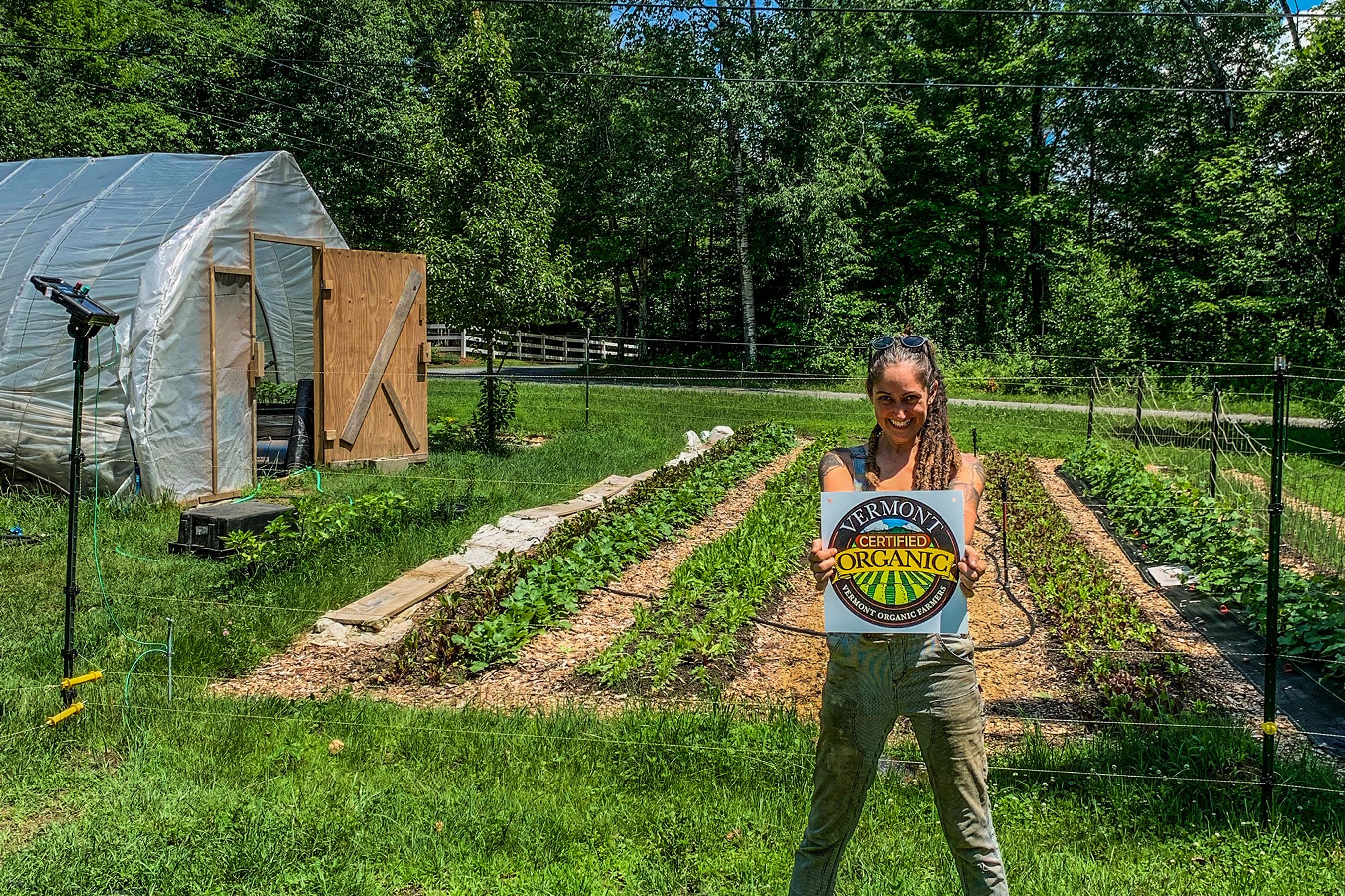 Journey Farmer Kirsten Tyler of Rocky Hill Farm stands proudly in front of her veggie fields holding her VOF organic certification seal
