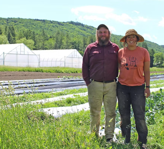 Farmers Gabrielle Tuite and Henry Webb of Old Road Farm stand smiling with their lush organic farm fields and high tunnels in the background