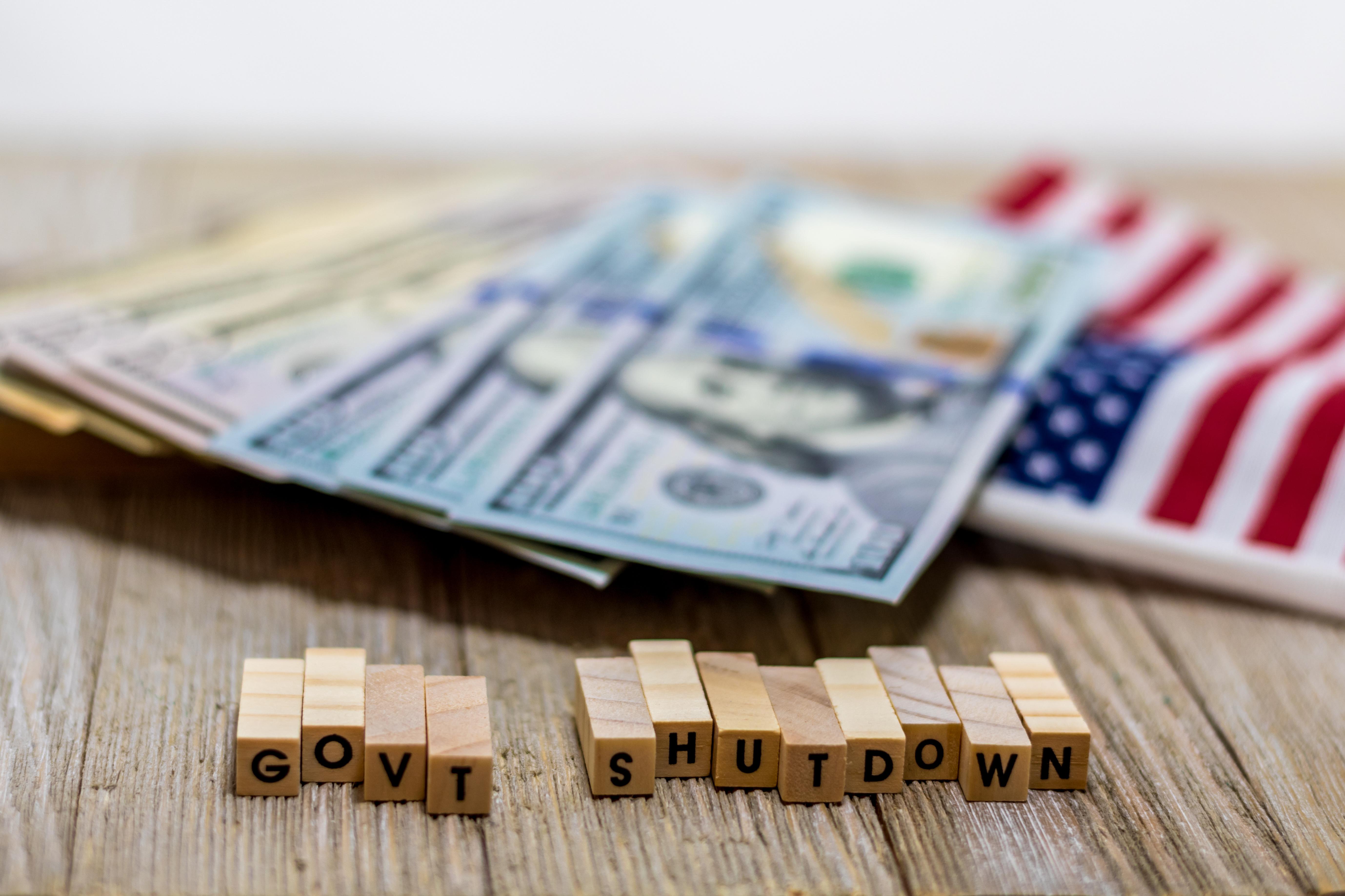 United States currency and an American Flag laying on a table in the background with small block letters reading "govt shutdown" in the foreground