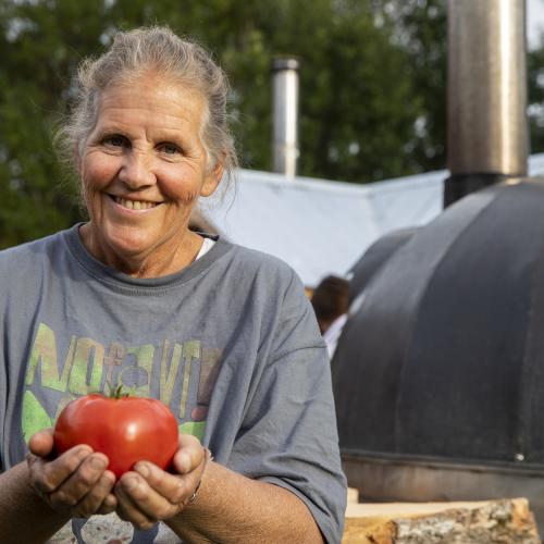 Enid Wonnacott stands outside in a NOFA-VT tee-shirt holding a tomato out to the camera and smiling. A woodfire pizza oven is in the background.