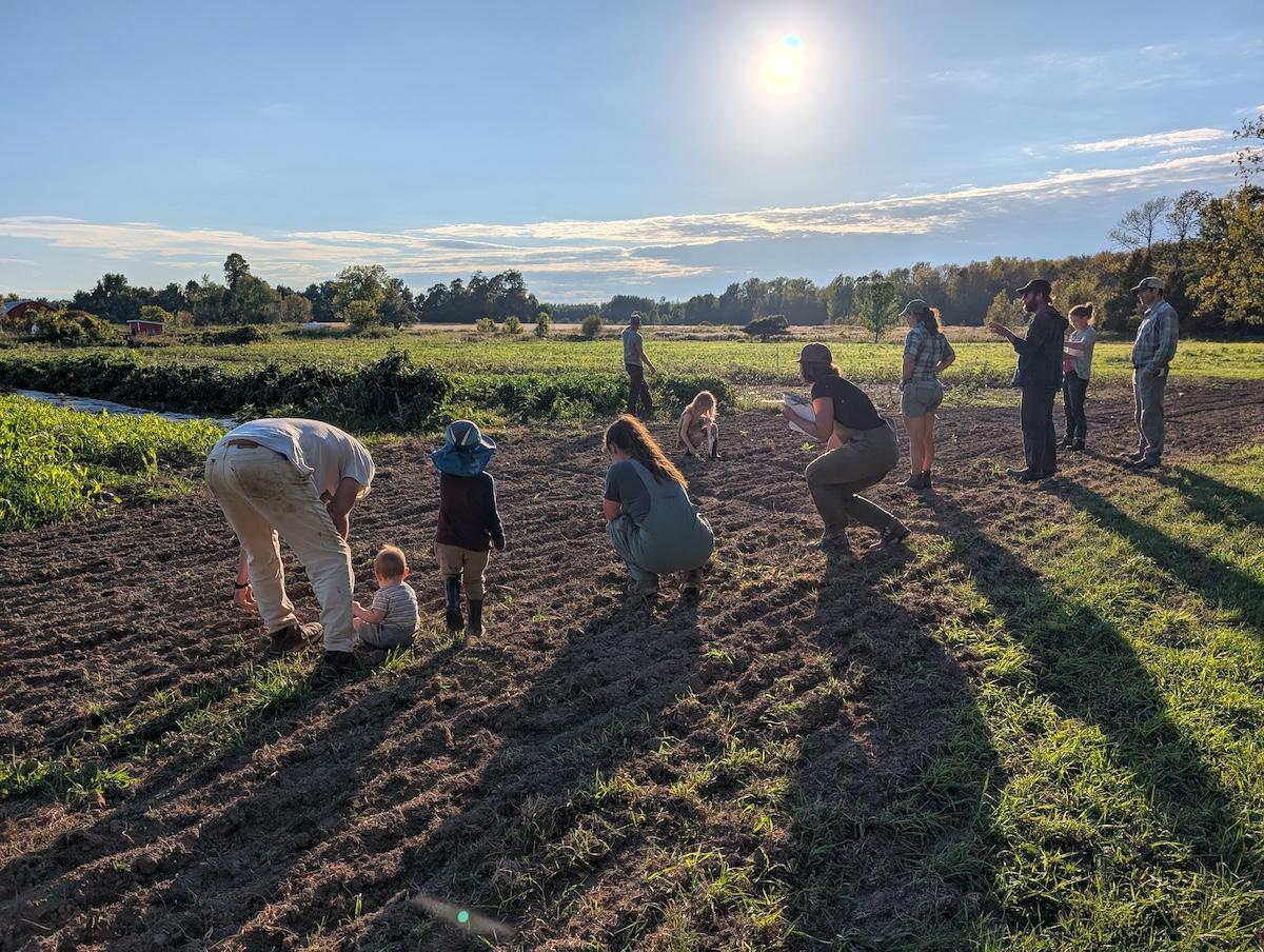 A group of about 10 adults and children look at the soil in a farm field.