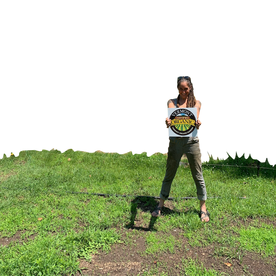 A person stands in a field, smiling and holding a Vermont Organic Farmers sign