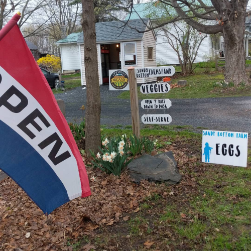 photo of an business "open flag", with a sign that lists products for sale.  There is a small farmstand shed in the background