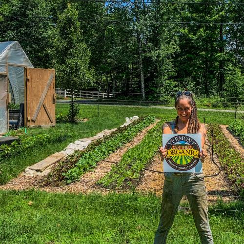 Kirsten Tyler of Rocky Hill Farm proudly beams while holding her VOF certification seal. She's standing in front of one of her organic veggie plots.