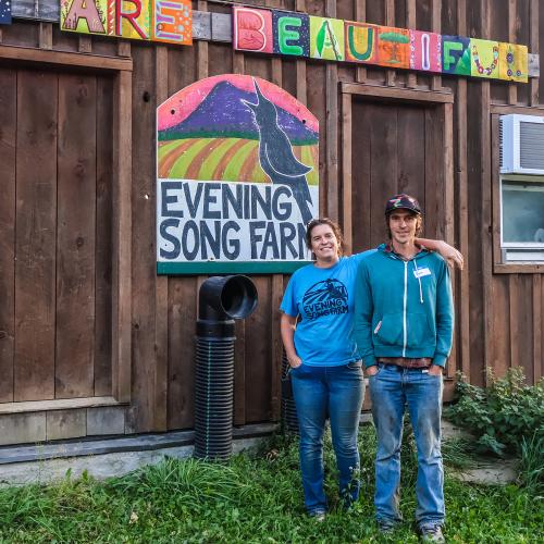 Ryan and Kara of Evening Song Farm stand in front of their barn with a colorful painting of their logo and a sign reading "you are beautiful"