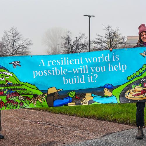 Two NOFA-VT staff members hold opposite ends of a colorful banner that reads "A resilient world is possible - will you help build it?"